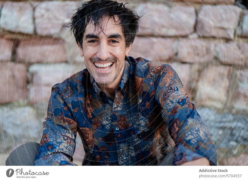 Smiling man with wet hair in patterned shirt smile happy cheerful genuine sitting stone wall casual joy excitement positive content adult male daylight outdoor