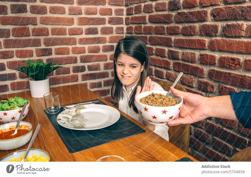 A young girl looks skeptically at a bowl of food being offered to her at the family dining table against a brick wall background weekend home picky eater meal