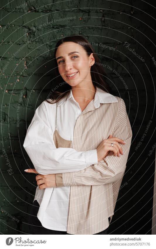 Attractive confidence young stylish woman in trendy shirt looks at camera, smiles, stands with crossed arms on brick wall. Beauty, fashion, self-expression, creativity, individuality concept