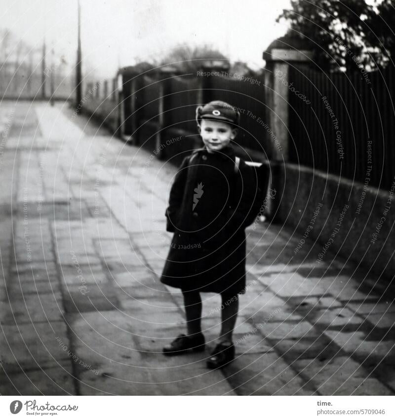 on the way to school Boy (child) Stand then Former portrait garments off Sidewalk Coat Satchel Wall (barrier) Street Looking into the camera Cap Contrast