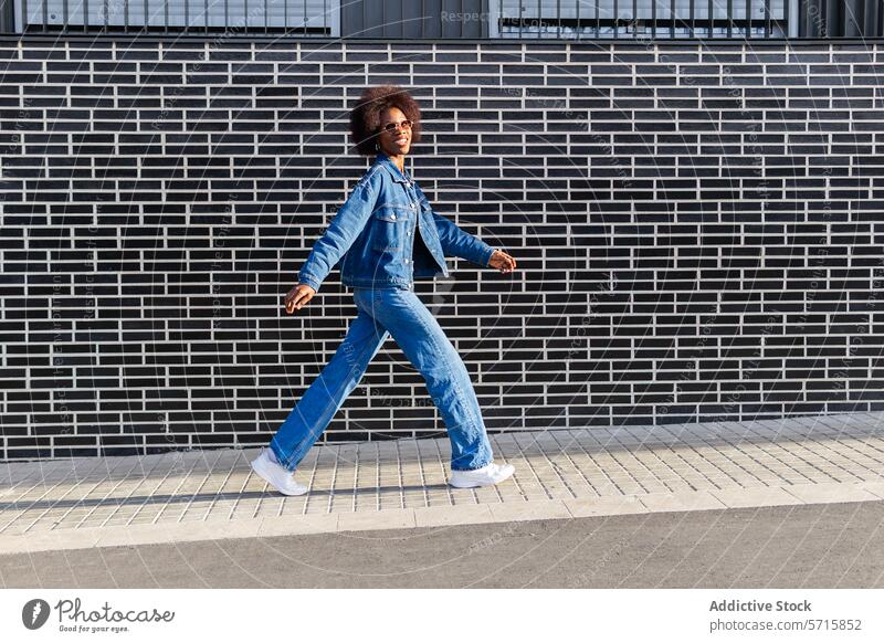 A confident African American woman in full denim strides forward, her dynamic pose set against the graphic lines of a brick wall style full body outfit fashion