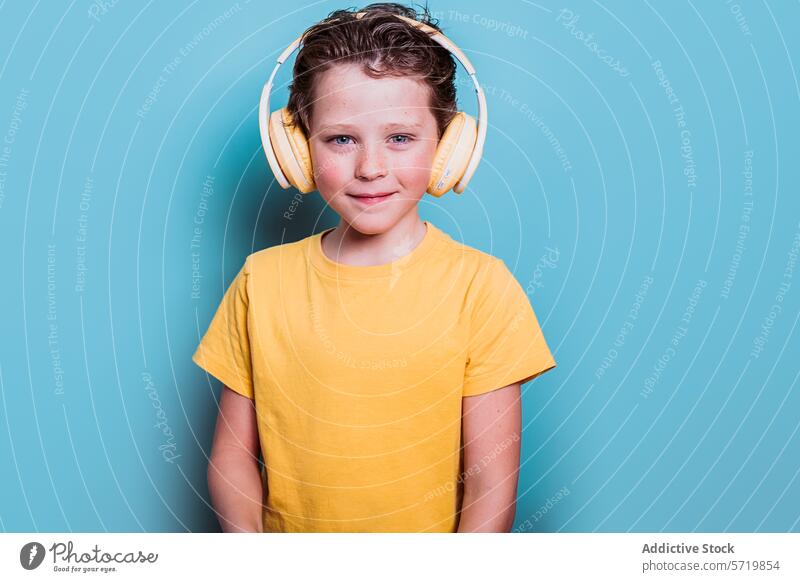 A cheerful school boy wearing a yellow t-shirt and headphones stands against a blue background, smiling warmly smile child student happy audio technology