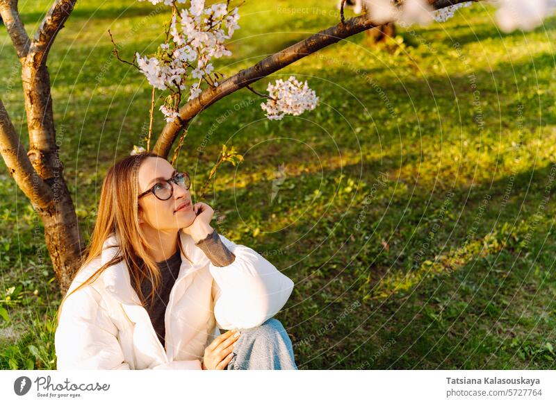 Beautiful young woman in white jacket and glasses sitting near blooming tree garden nature cherry outdoor blossom beauty eyeglasses park beautiful long spring