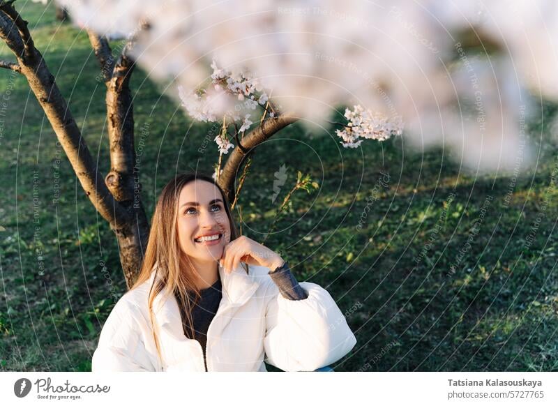 Beautiful woman in a white jacket sits under blooming tree in city park in spring young garden nature cherry outdoor blossom beauty beautiful long trees scenic