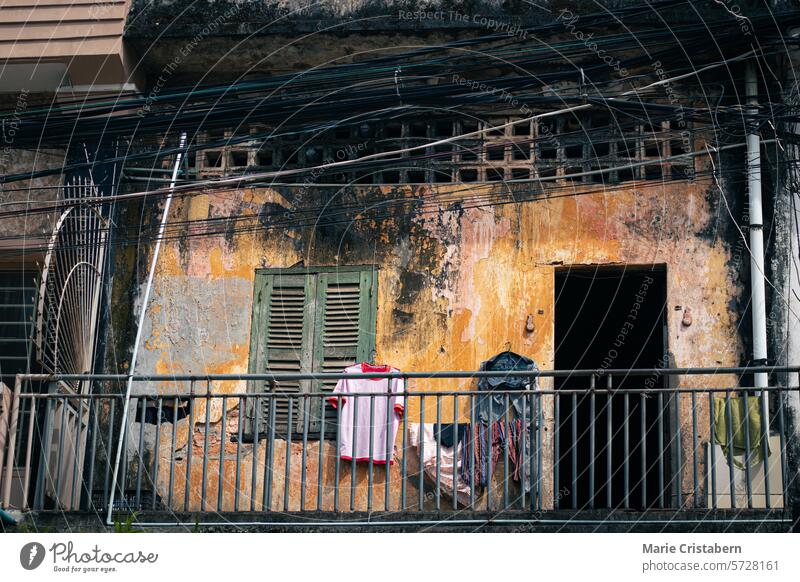 Weathered balcony of an old rustic colonial building with laundry hanging, green shutters, railing, and visible wires overhead showing a candid scene of the khmer daily life in Kampot, Cambodia