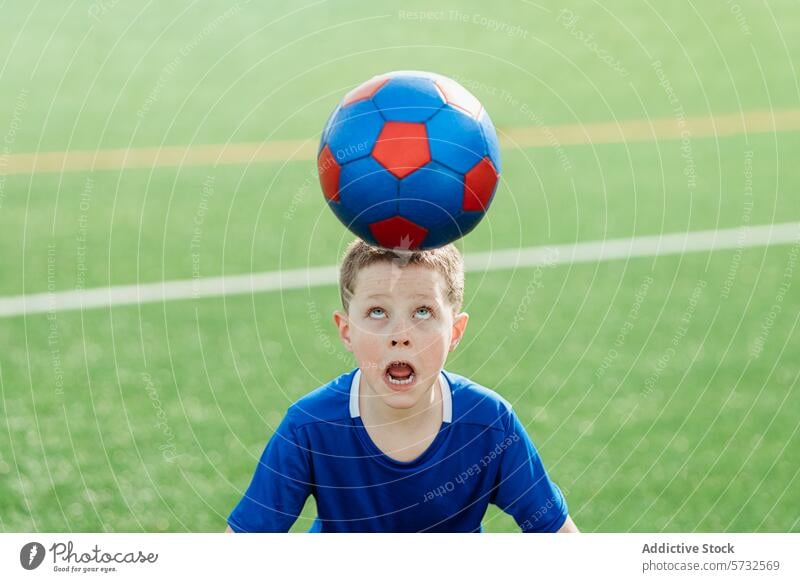 Young soccer player balancing ball on head boy balance concentrate jersey blue red young sport field grass outdoor game skill training childhood practice