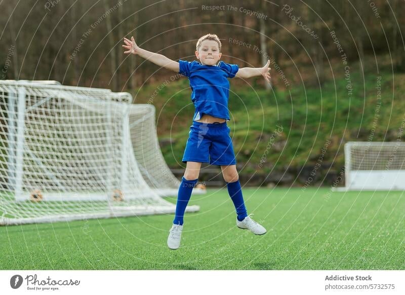 Young soccer player celebrating on the field boy sport celebration joy football young jump uniform outdoor goal net arms stretched grass green blue kit happy