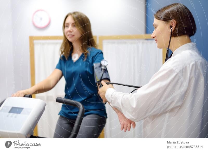Female doctor cardiologist measures pressure with patient during bicycle exercise for examination cardiovascular system at medical office of clinic. Young woman training on bike simulator.