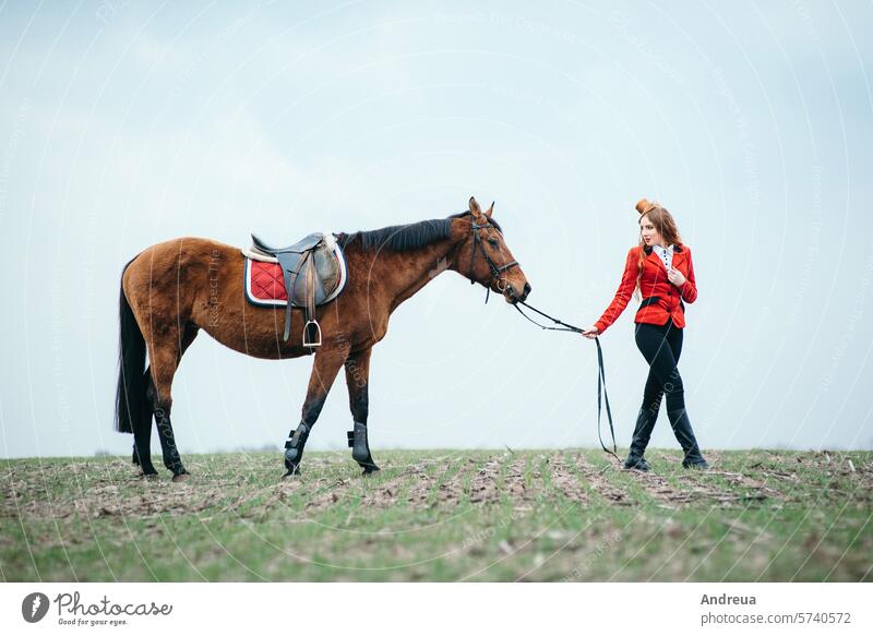red-haired jockey girl in a red cardigan and black high boots with a horse woman saddle ride summer nature equestrian training stallion rider lifestyle outdoor