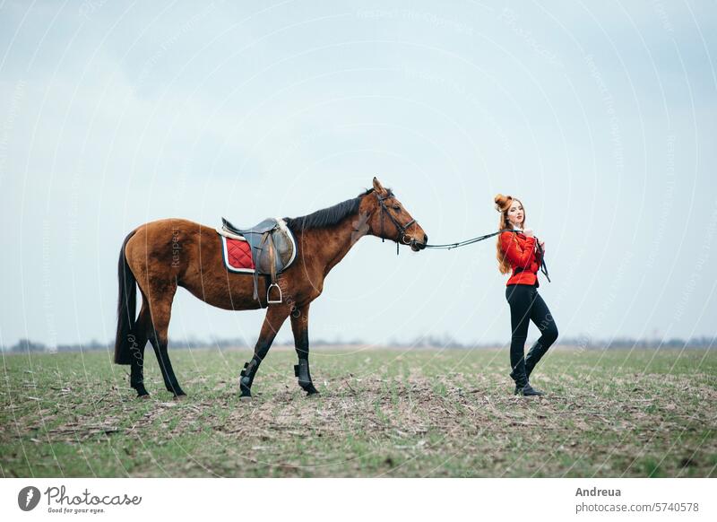 red-haired jockey girl in a red cardigan and black high boots with a horse woman saddle ride summer nature equestrian training stallion rider lifestyle outdoor