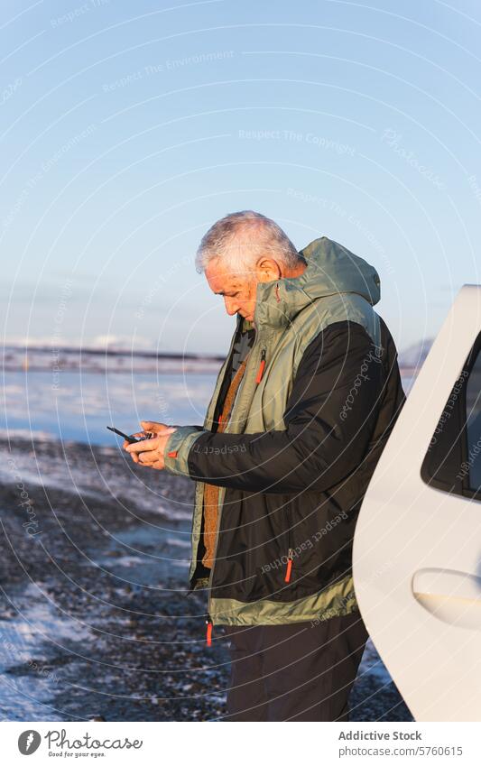 A senior traveler engages with his smartphone, standing by a vehicle on an icy terrain under the soft light of the Icelandic winter sun man technology