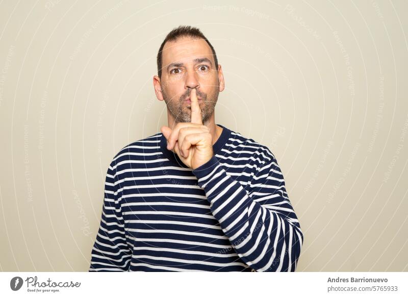 Hispanic man with a beard in his 40s with a striped sweater putting his index finger to his mouth asking for silence, he is keeping a secret. Isolated on beige studio background.