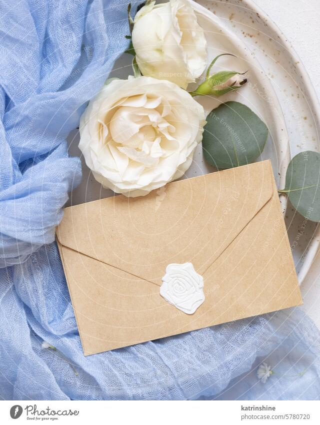 Envelope near blue tulle fabric knot and cream roses on plates top view copy space, wedding mockup envelope sealed romantic flowers white silk spring