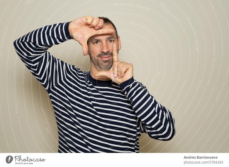 A happy bearded man blinks his eyes and looks through a frame formed by his hands. He makes a frame with his fingers, isolated on beige background with copy space for text. People concept.