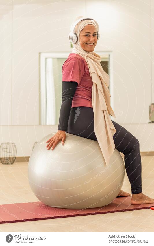 Muslim woman smiling on a fitness ball before yoga class muslim gym workout headphones preparation active lifestyle health exercise ethnic attire hijab