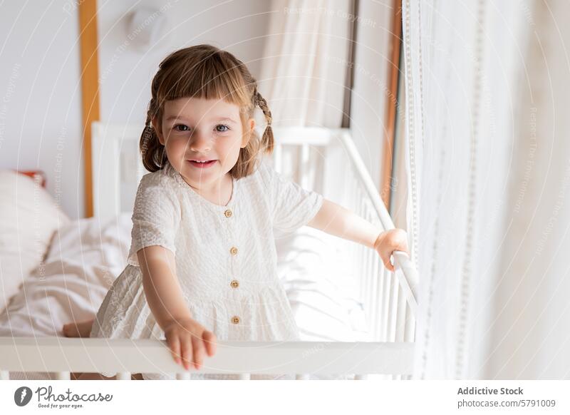 Adorable toddler in white dress standing by crib girl bedroom child smile braids sunny cheerful innocence cute adorable little youngster playful joy happiness