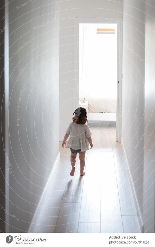 Toddler Girl Walking Towards Light in Hallway toddler girl walking barefoot hallway door open natural light bright home indoor child explore curiosity back view