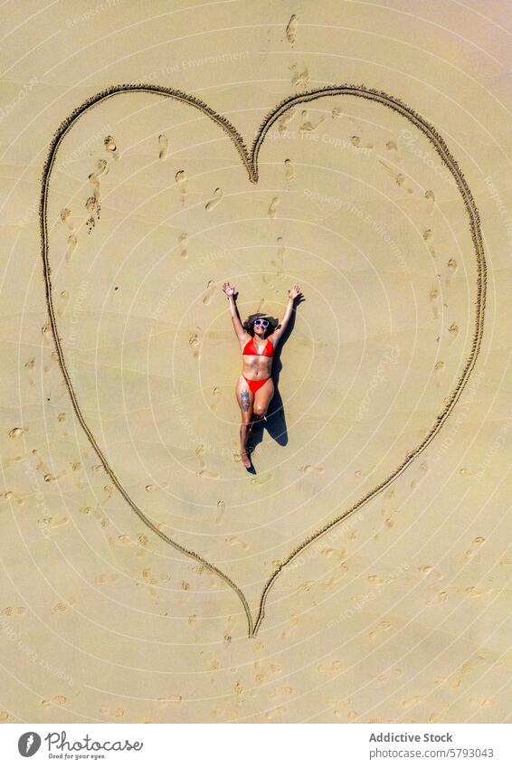 Joyful beach day with a heart in the sand woman aerial view sunny seaside lying down sand drawing shoreline swimwear vacation holiday sunglasses summer