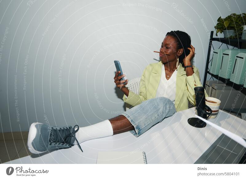 Businesswoman sitting with foot on office desk and taking selfie holding pencil between lips and nose Selfie fun photograph funny posing work playful