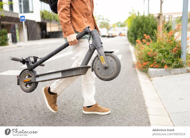 Man carrying an electric scooter on a city street man transport urban sidewalk casual attire convenient modern greenery shift environment portable mobility