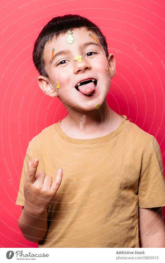 Playful boy with stickers on face making silly face playful tongue gesture pink background cheerful young child kid fruit vibrant fun school schoolkid male