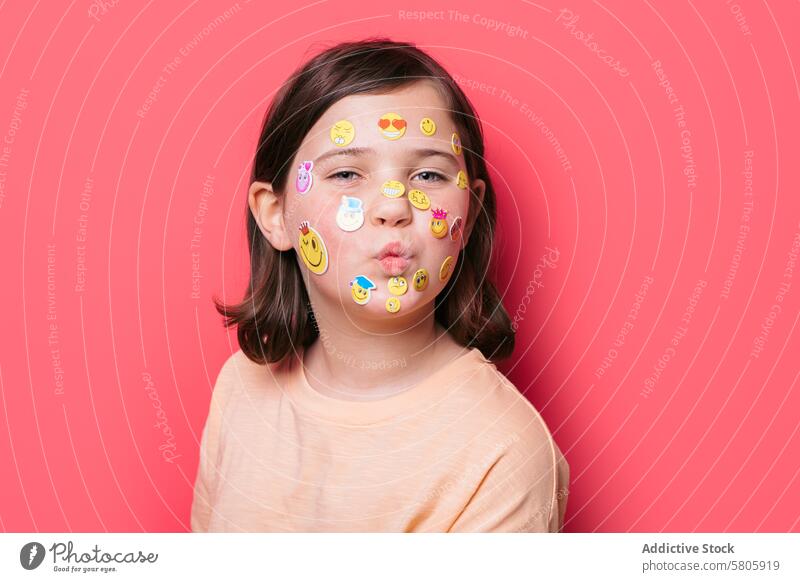 Young girl with emoji stickers on face making pout child decoration fun playful expression cute school kid emotion casual short hair adhesive pink background