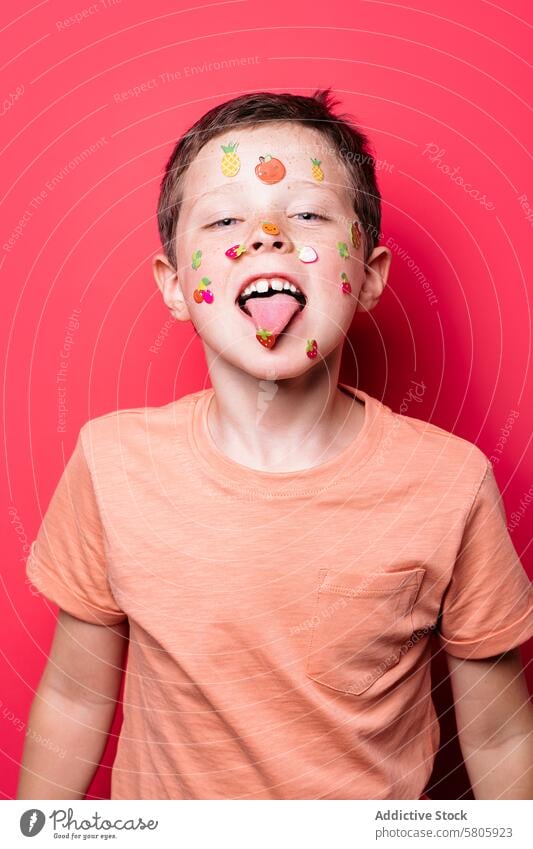 Playful boy with sticker-covered face on red background child playful stickers tongue school kid looking at camera cheerful fun vibrant colorful expression joy