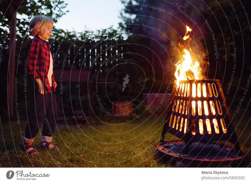 Cute little boy is burning a bonfire on a summer evening in the backyard. Child puts firewood in a fire bowl by tongs. Summer holidays for kids in village.