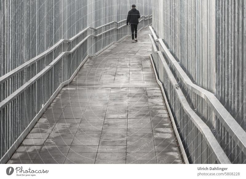 A person dressed in black walks along a paved path bordered on the left and right by steel struts, with handrails pointing backwards in perspective. off