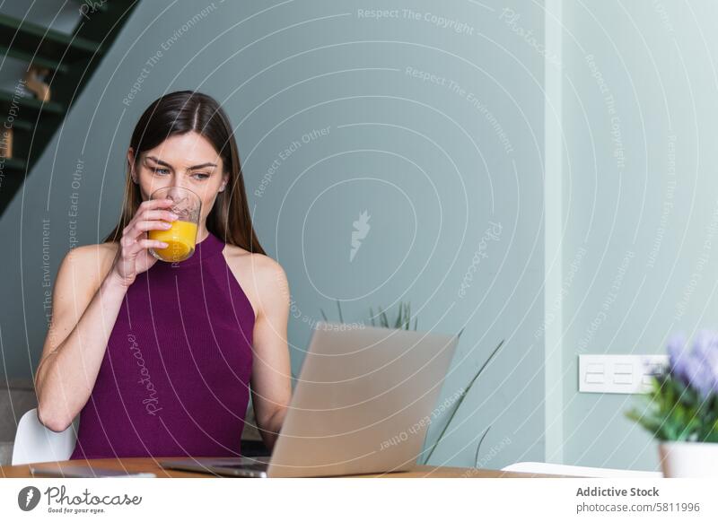 Cheerful woman browsing laptop in living room using toothy smile happy gadget juice drink cheerful netbook casual glass orange attractive surfing work beverage