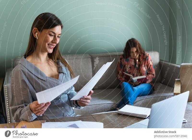 Cheerful woman reading documents and sitting at desk with laptop women cheerful cowork office colleague toothy smile paperwork earbuds book positive casual