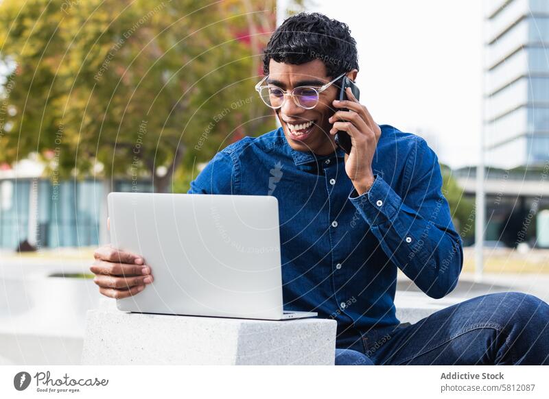 Happy ethnic businessman freelancer working on laptop and talking on mobile phone in city employee using gadget speak device phone call netbook conversation