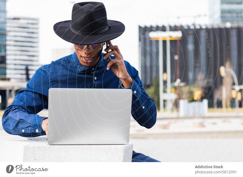 Serious ethnic businessman freelancer working on laptop and talking on mobile phone in city employee thoughtful using gadget serious speak device phone call