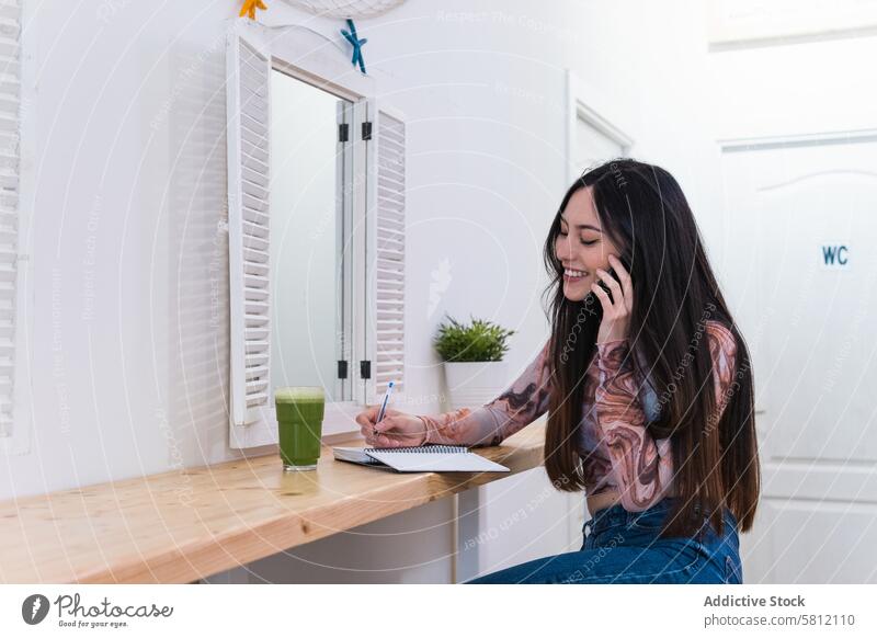 Woman taking notes in notebook and speaking on mobile phone sitting in bar with healthy beverage woman green smoothie take note phone call write enjoy chill