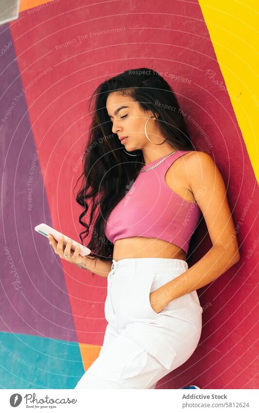 Latin woman using smartphone leaning on a colorful wall young summer female outdoors people lifestyle joyful spring enjoyment hair city cool stylish outfit