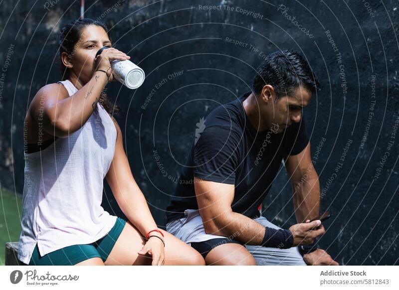 Ethnic woman drinking water near athlete using smartphone training thirst break sportswoman sportsman couple internet cellphone device activewear ethnic