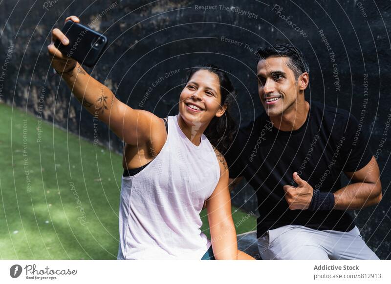 Smiling ethnic couple taking selfie on smartphone during training sportswoman sportsman using thumb up gesture break workout positive approve device happy fit