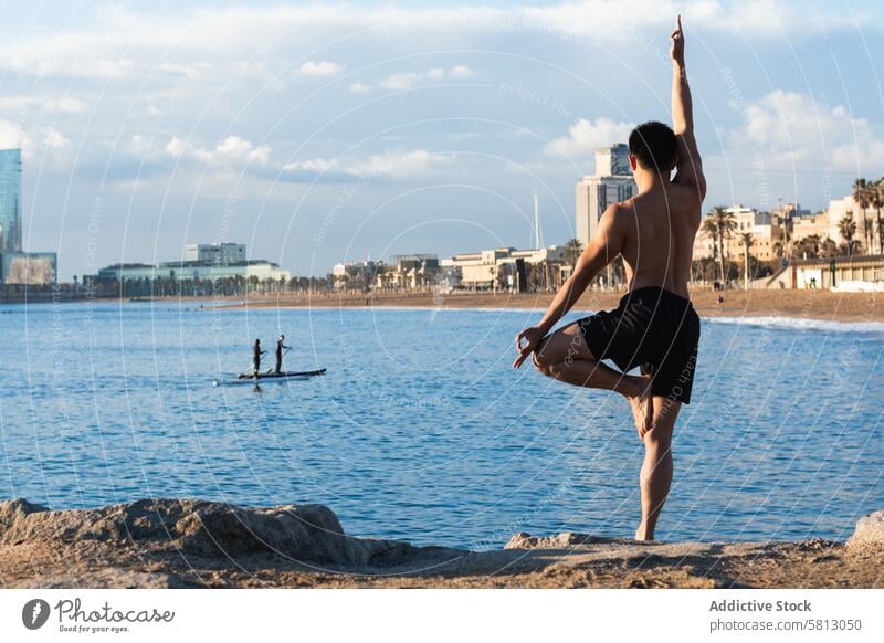 Shirtless man balancing in yoga pose on beach tree pose vriksasana practice balance stand male shirtless mudra gesture lifestyle wellness wellbeing vitality