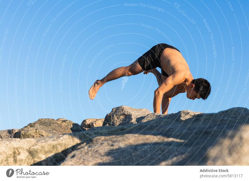 Ethnic man doing arm balance yoga pose on stone asana flying pigeon eka pada galavasana advanced practice handstand male shirtless lifestyle wellness wellbeing