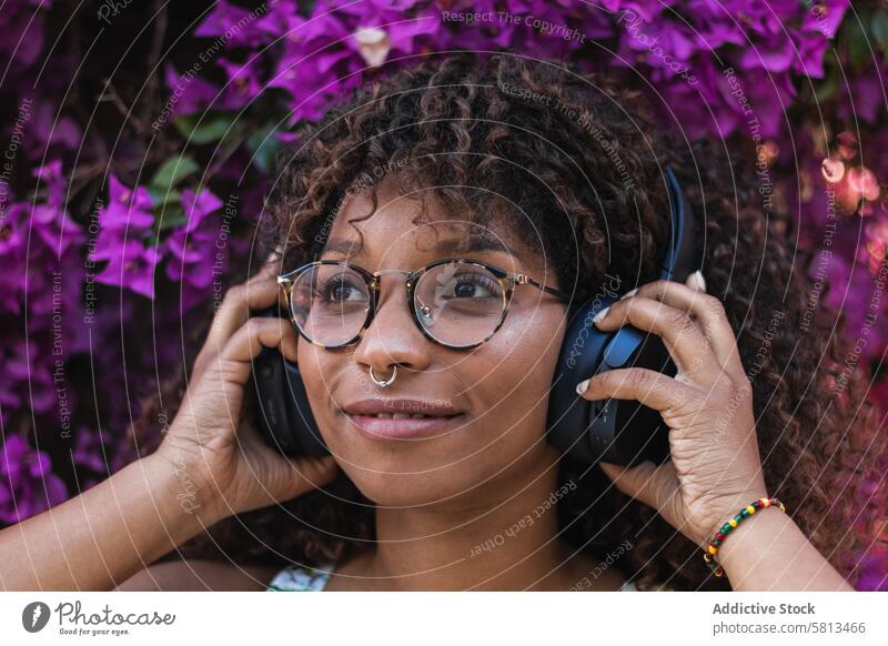 Black woman listening to music near flowers bush garden headphones smile style young female glasses piercing ethnic black african american song joy trendy