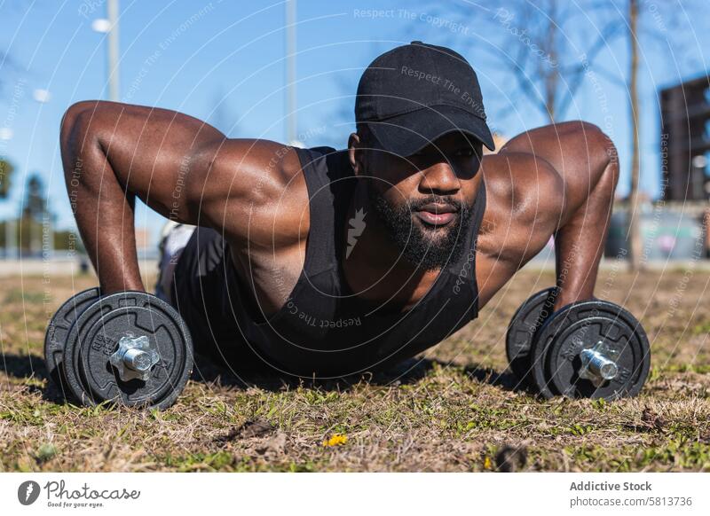 Strong African American sportsman pushing up on dumbbells in town push up fitness training muscle workout activity lawn bicep grass athlete exercise cap