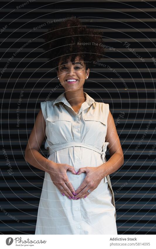Smiling black woman showing heart gesture on pregnant tummy maternal happy expect belly anticipate prenatal female positive pregnancy glad abdomen optimist