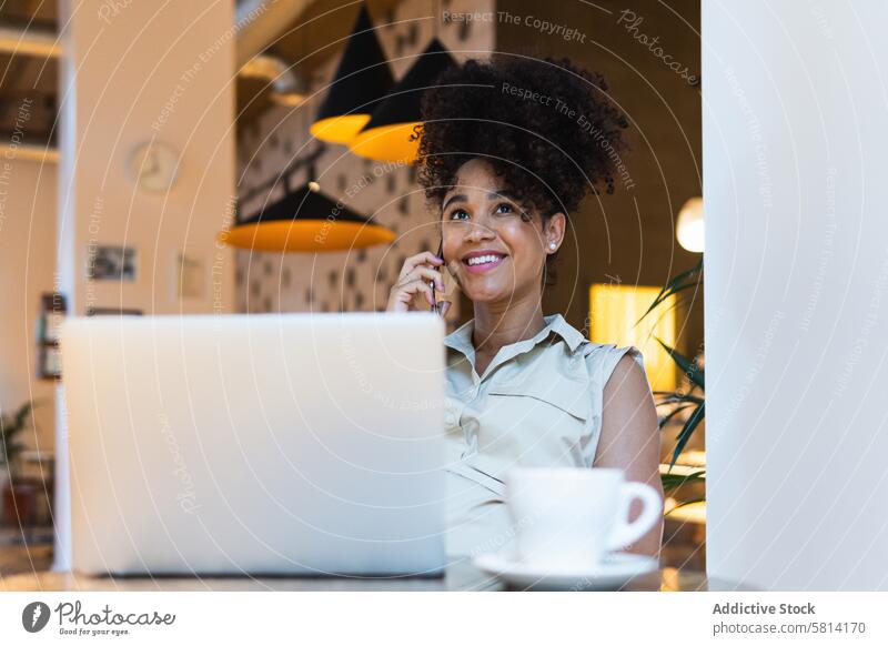 Smiling black woman talking on smartphone at table with laptop using positive speak phone call happy connection freelance female conversation coffee communicate