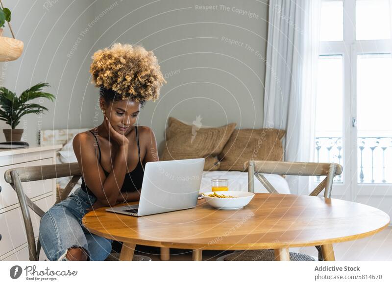 Black woman with laptop sitting on chair during breakfast watch work using remote online busy morning female device browsing gadget home connection serious