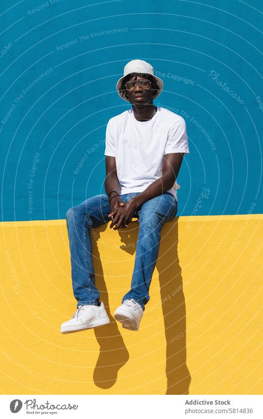 Happy ethnic man sitting on colorful wall cheerful happy summer sunlight optimist bright casual hat sunglasses young black male african american carefree