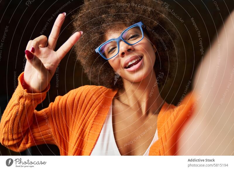 a young woman with afro hair taking a self-portrait with glasses and a victory gesture happy dark background fashionable beauty female person face attractive