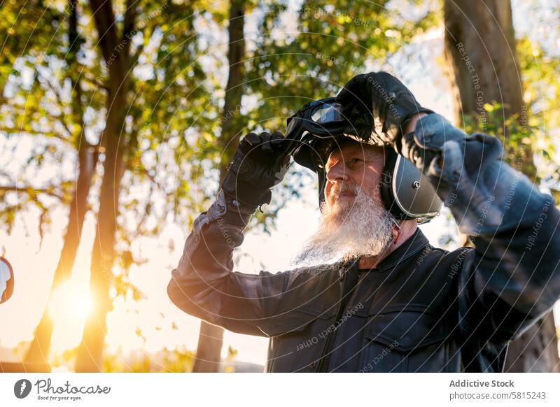 Senior Biker Adjusting Goggles Outdoors at Sunset senior male biker goggles protective gear outdoors sunset white beard mature looking away helmet motorcycle