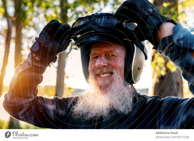 Smiling Senior Biker with Goggles Enjoying Sunset senior man biker motorcycle helmet goggles smiling outdoors sunset looking at camera beard white happiness