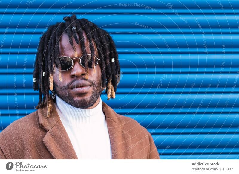 Black man in trendy sunglasses in city dreadlocks style urban cool hairstyle accessory male ethnic black african american blue wall fashion lifestyle confident