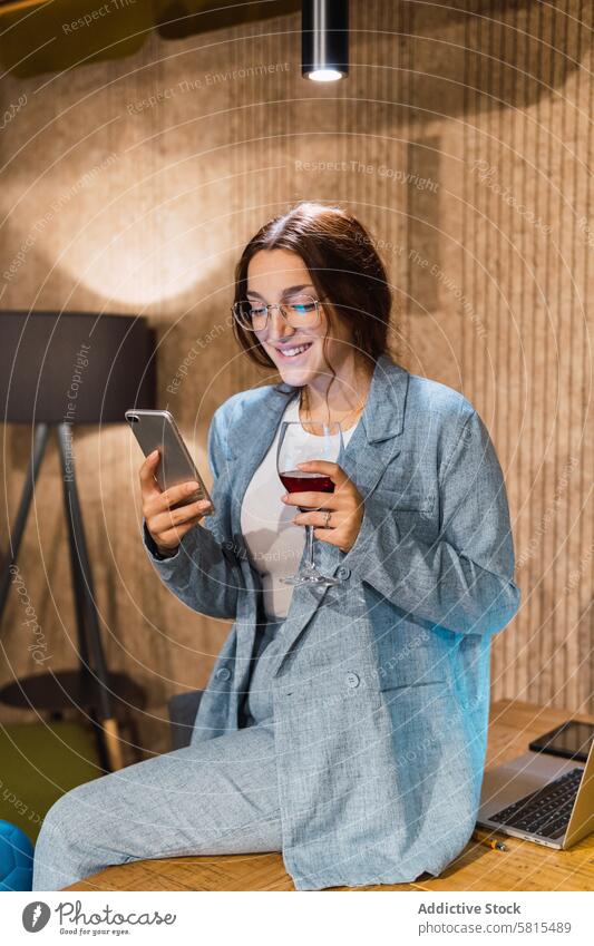 Young businesswoman with wine using smartphone break workplace smile red wine social media freelance table female young laptop online gadget internet surfing
